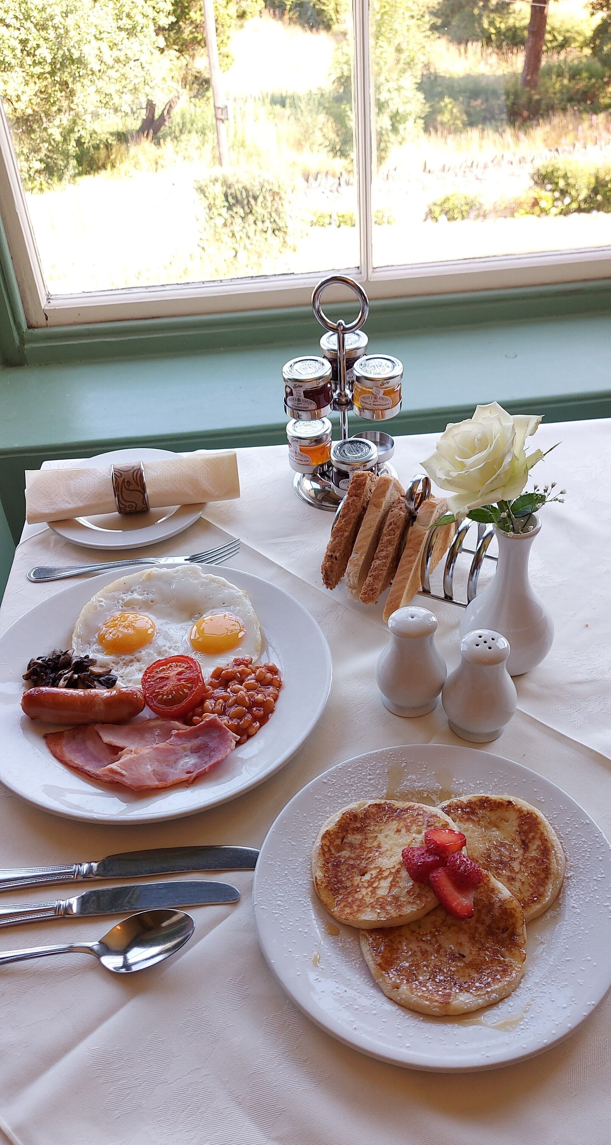 A breakfast table with plates of eggs, bacon, beans, and toast, a vase with a white rose, salt and pepper shakers, and a window view of greenery.