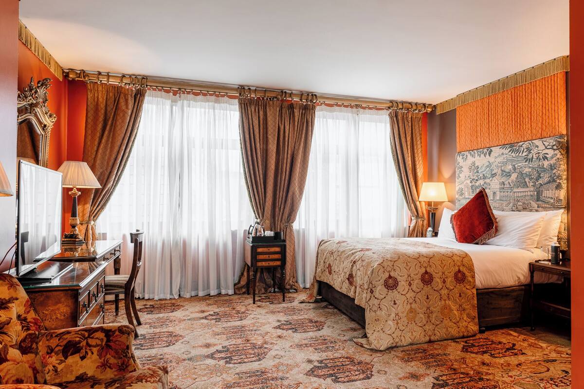 A hotel room with a large bed, a desk with a lamp, a chair, and a carpeted floor.