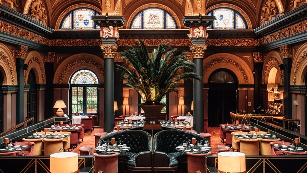 A grand hotel lobby with ornate architecture, a central seating area, and multiple dining tables.