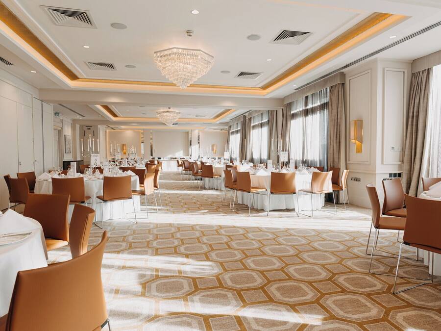 Un amplio salón de banquetes con alfombra de diseño hexagonal, mesas blancas y sillas marrones.