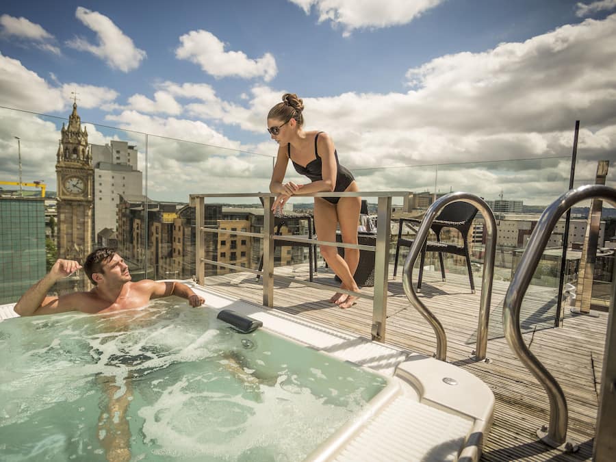 A rooftop pool area with a hot tub, metal railings, and a view of a clock tower in the background.
