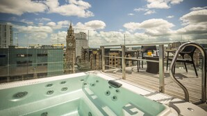 A rooftop terrace with a hot tub, outdoor seating, and a view of a clock tower and city buildings.