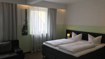 Habitación de hotel con cama, una silla, una mesita de noche con lámpara y una ventana con cortinas translúcidas.