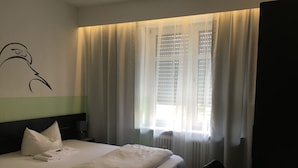 Un lit bien fait, avec des oreillers et une couverture pliée, une table de chevet avec une lampe, et une fenêtre ornée de rideaux légers.