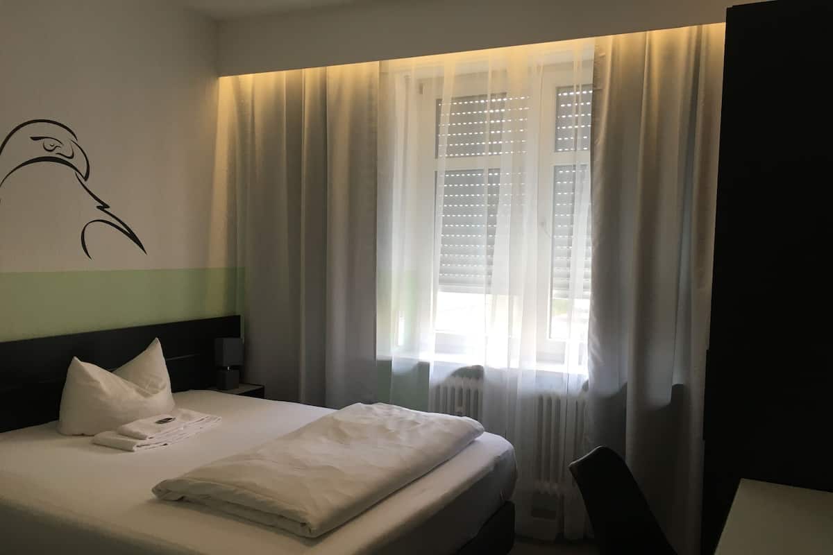 A neatly made bed with pillows and a folded blanket, a bedside table with a lamp, and a window with sheer curtains.