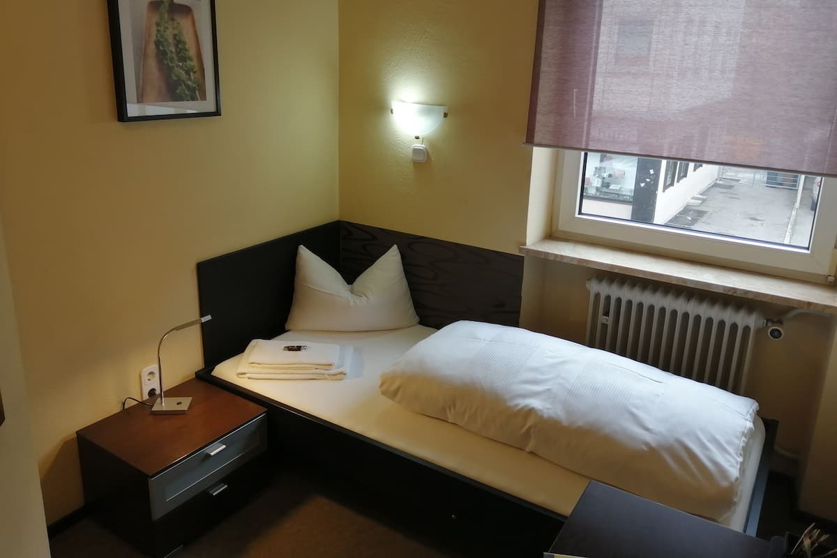 A small, neatly arranged bedroom with a single bed, a bedside table with a lamp, a framed picture on the wall, and a window with a view of the street.