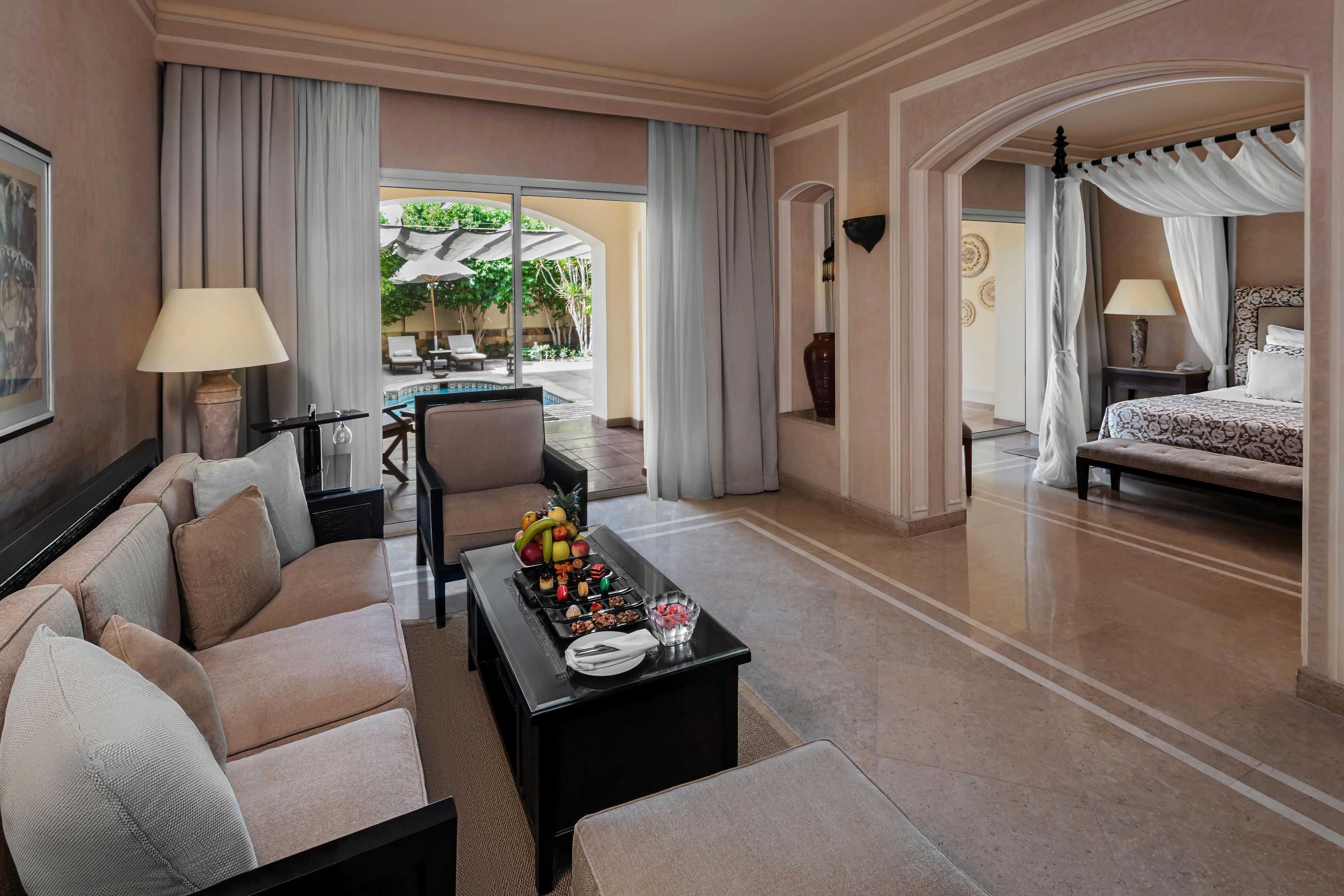 A hotel room with a sofa, a coffee table with fruit, a bed with a canopy, and a view of an outdoor area with umbrellas.