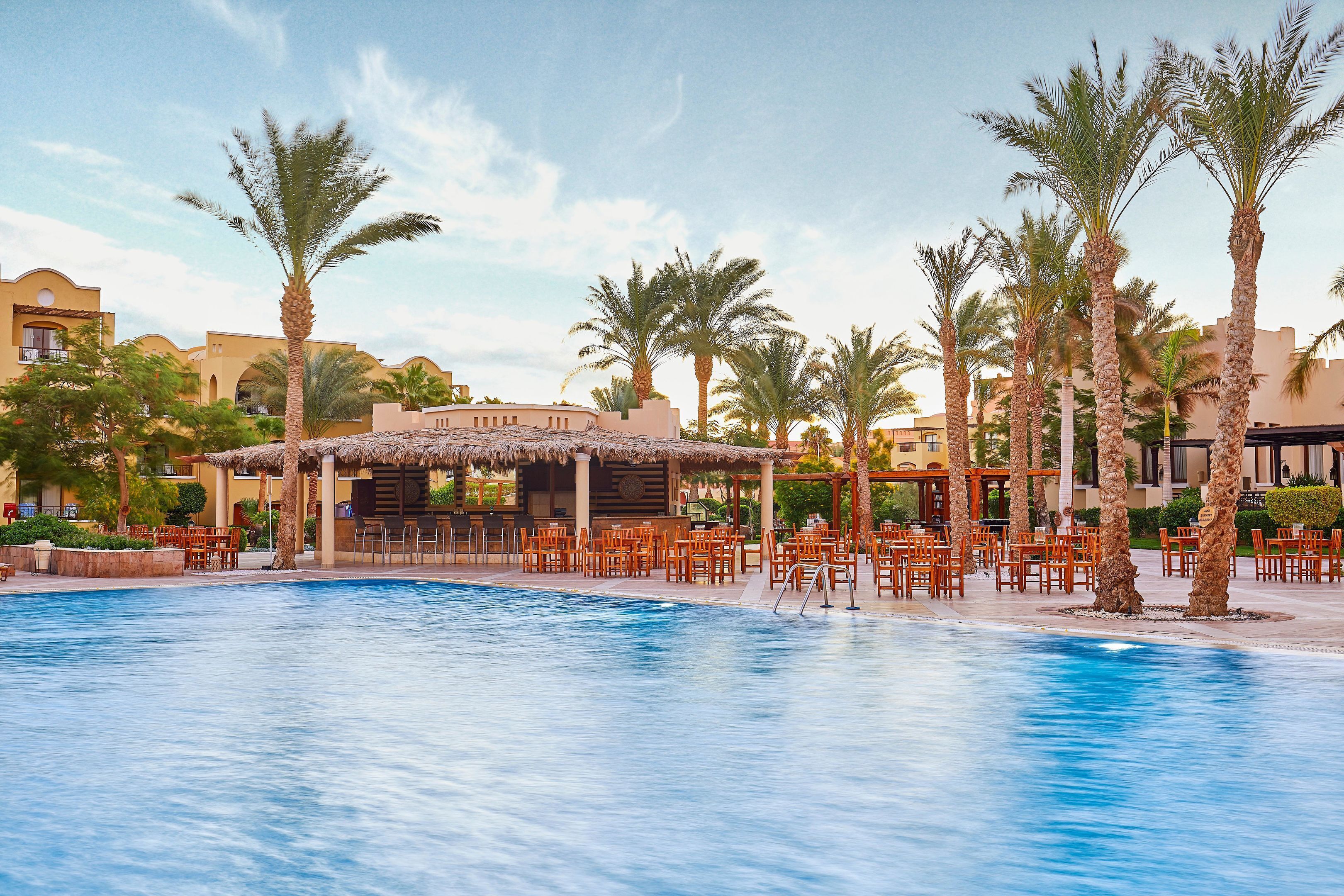 A pool area with palm trees, outdoor seating, and a thatched-roof structure.