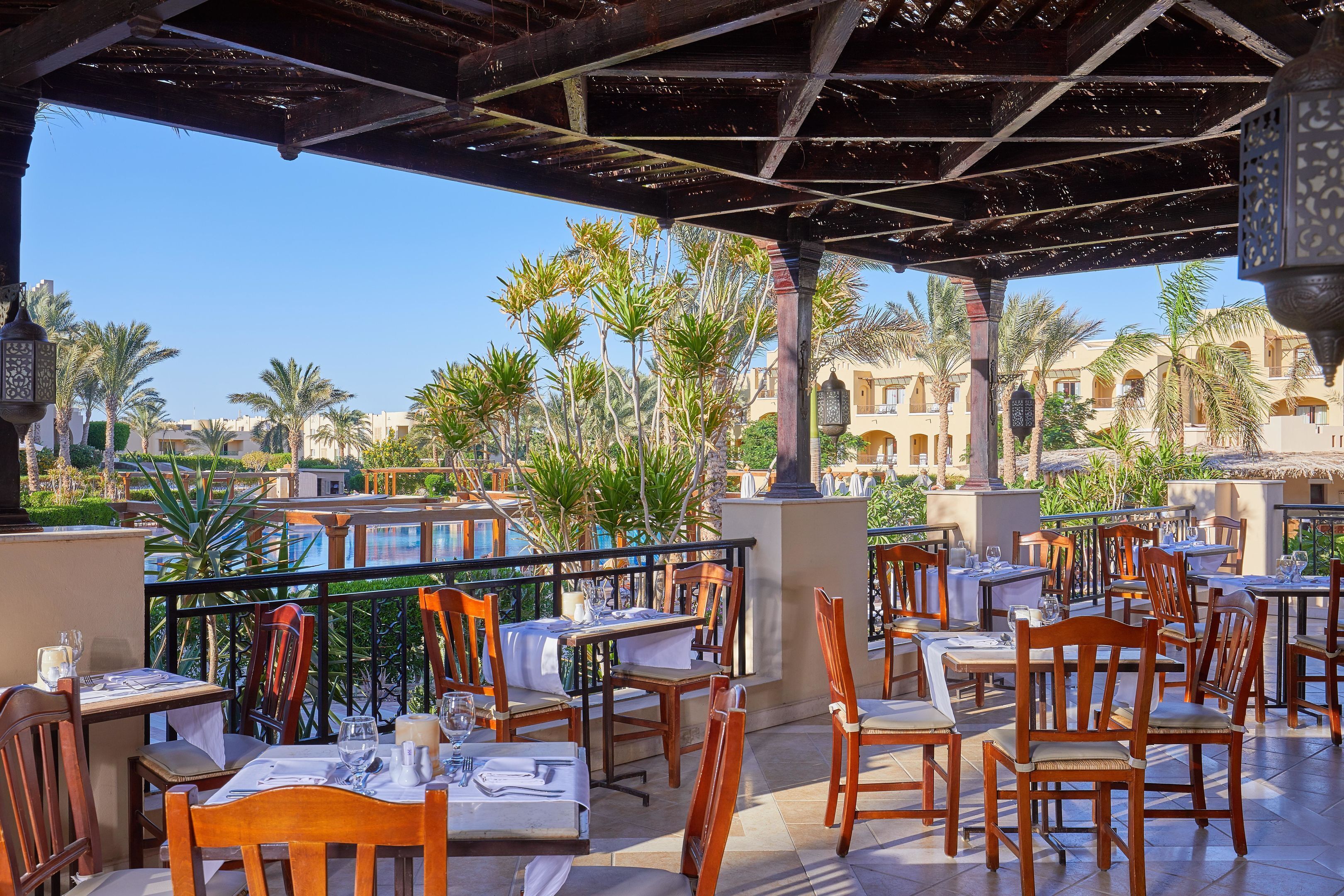 An outdoor restaurant with wooden tables and chairs, a covered area with a view of palm trees and buildings.
