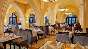 Um restaurante amplo com tetos abobadados e várias áreas de refeições. Mesas dispostas de forma organizada, com toalhas brancas e talheres e copos postos.