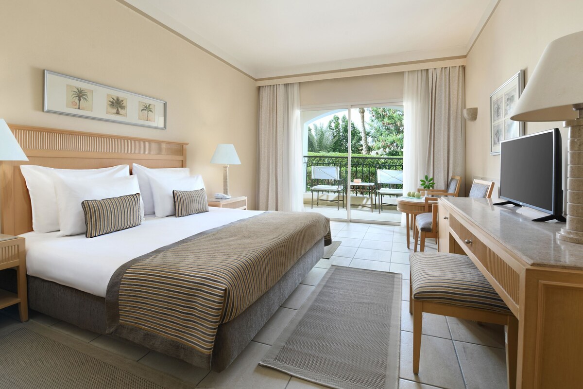 A hotel room with a large bed, a desk with a TV, a balcony with outdoor furniture, and framed artwork on the walls.