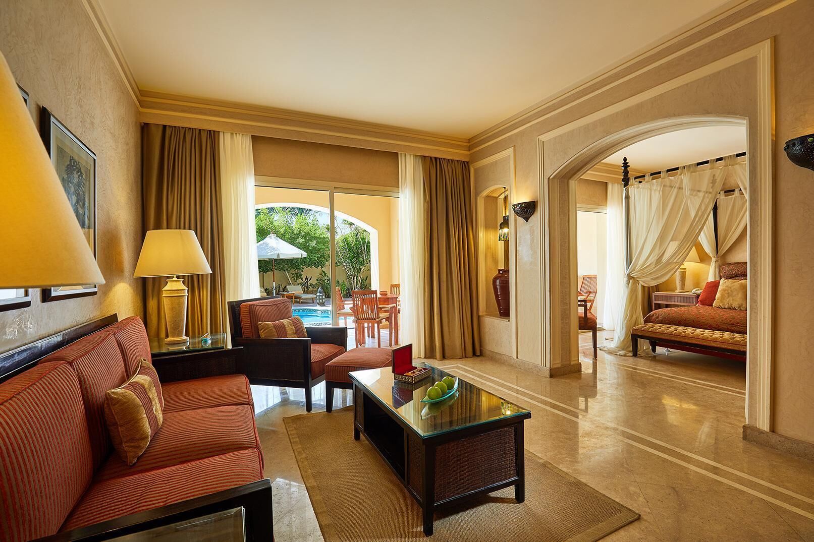 A hotel room with a sofa, armchair, dining table, and a view of an outdoor area with a pool and chairs.