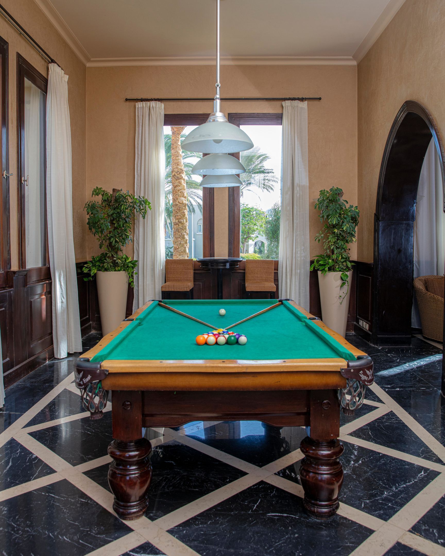 A pool table in a room with a marble floor, two potted plants, a chandelier, and a view through a window.