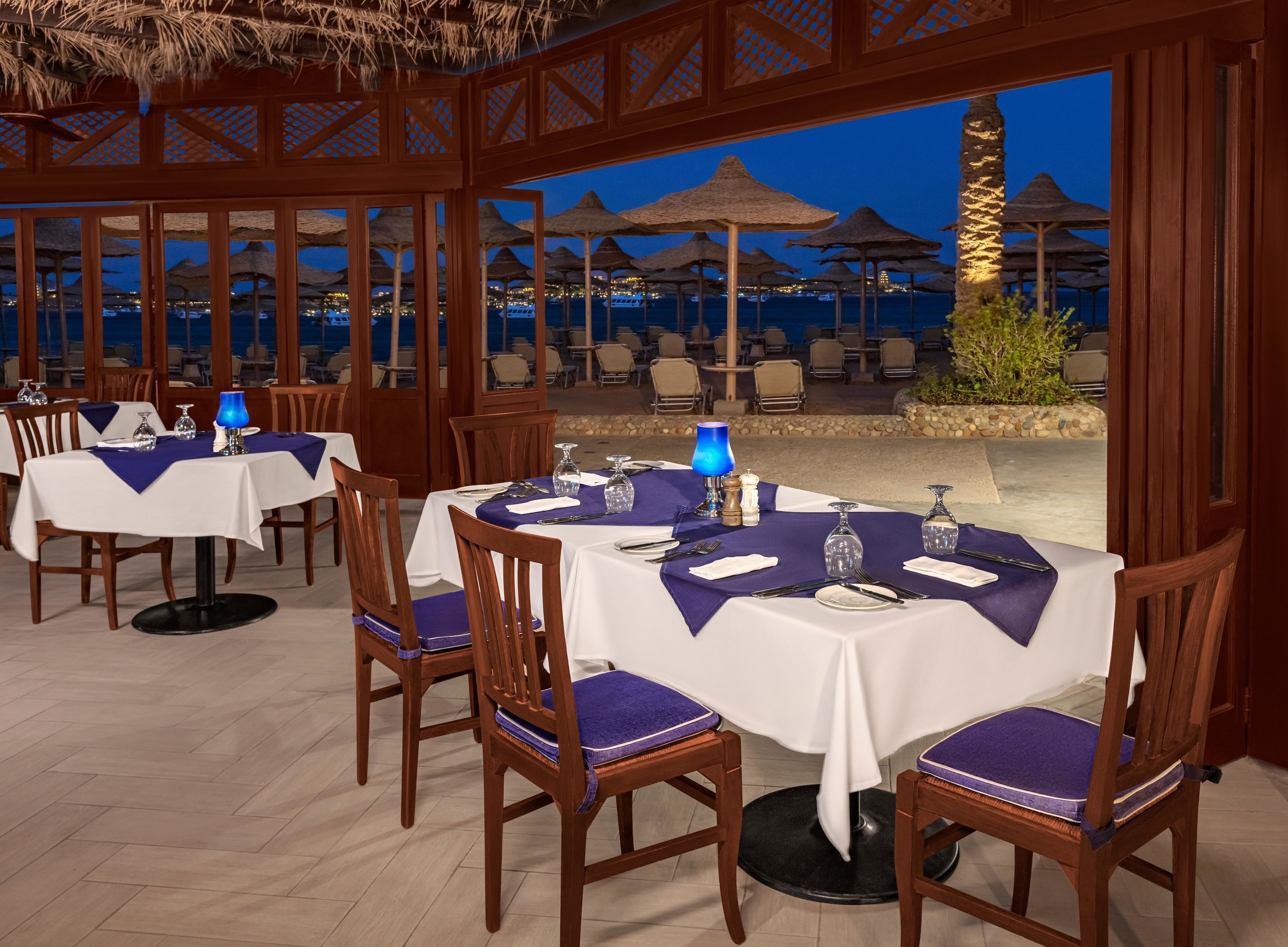 A restaurant with tables set for dining, chairs, and a view of the sea at night.