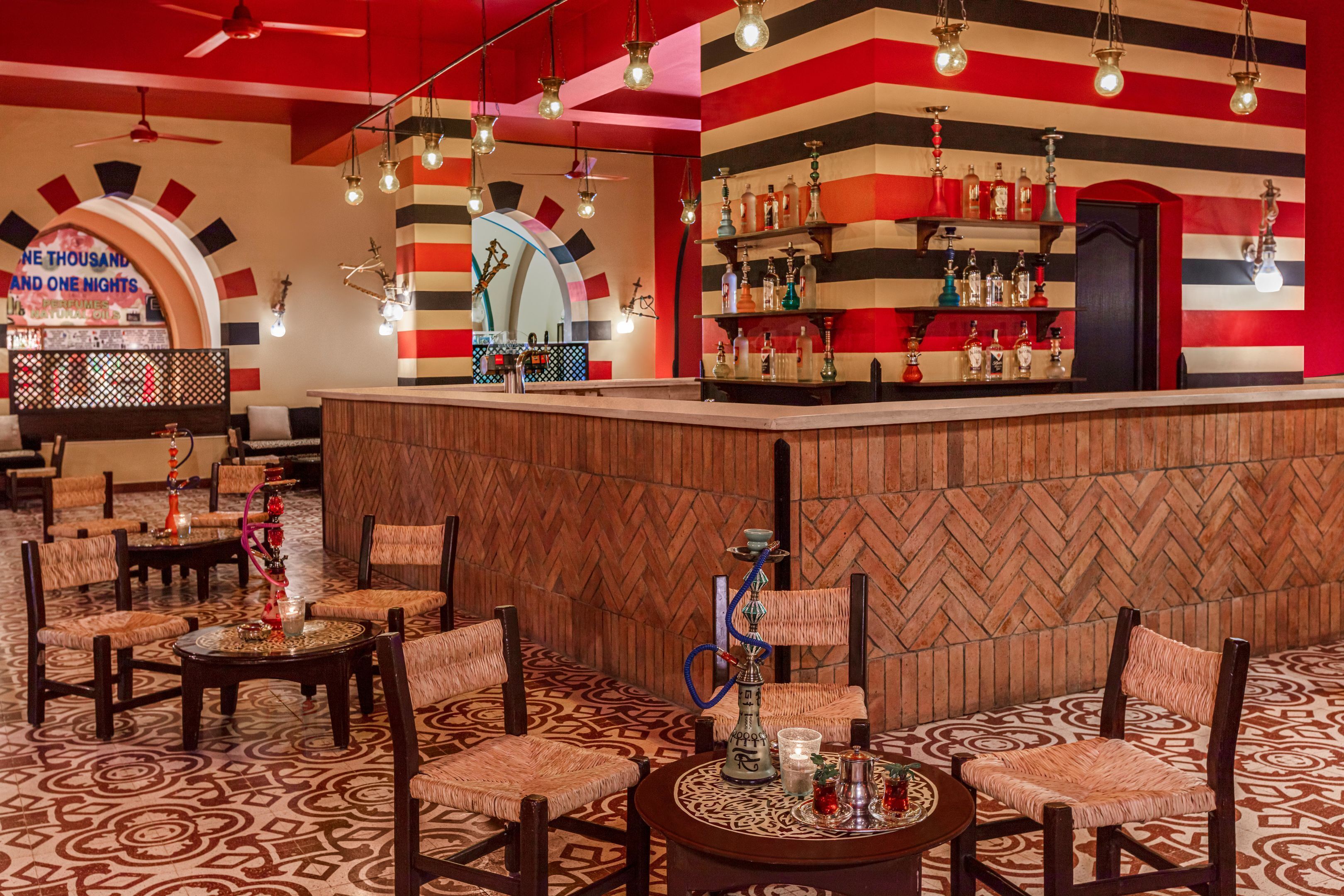 Zona de bar con pared de diseño en patrón espiga, mostrador de madera y área de asientos con suelo estampado y sillas tejidas.