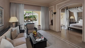 Quarto de hotel com sofá, mesa de centro com frutas, cama com dossel e vista para área externa com guarda-sóis.