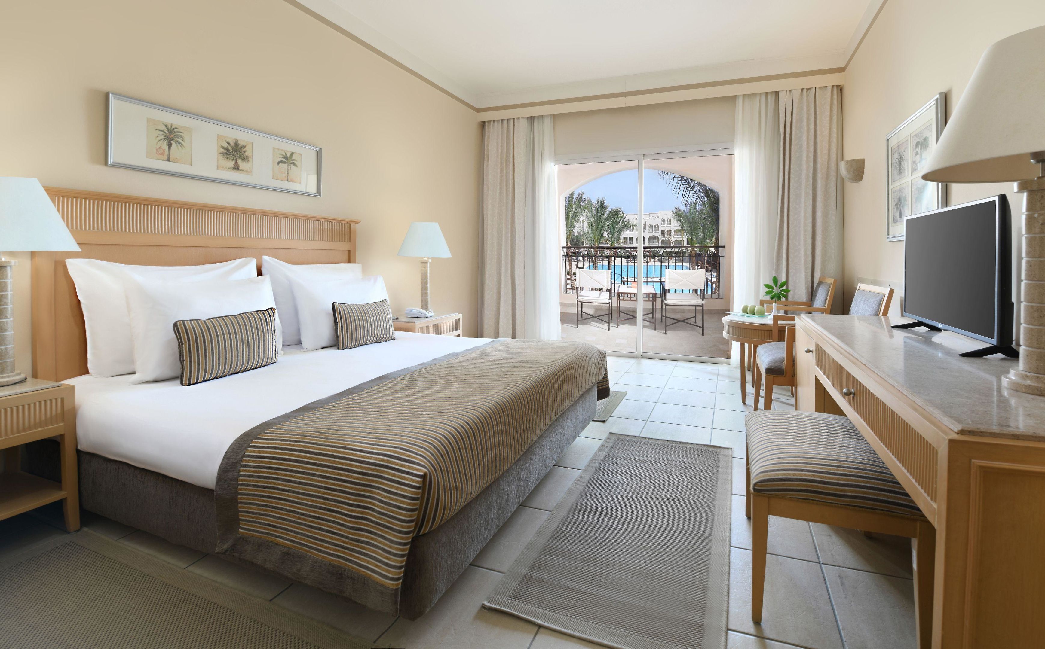 Habitación de hotel con cama, escritorio, televisión y balcón con vista a la piscina.
