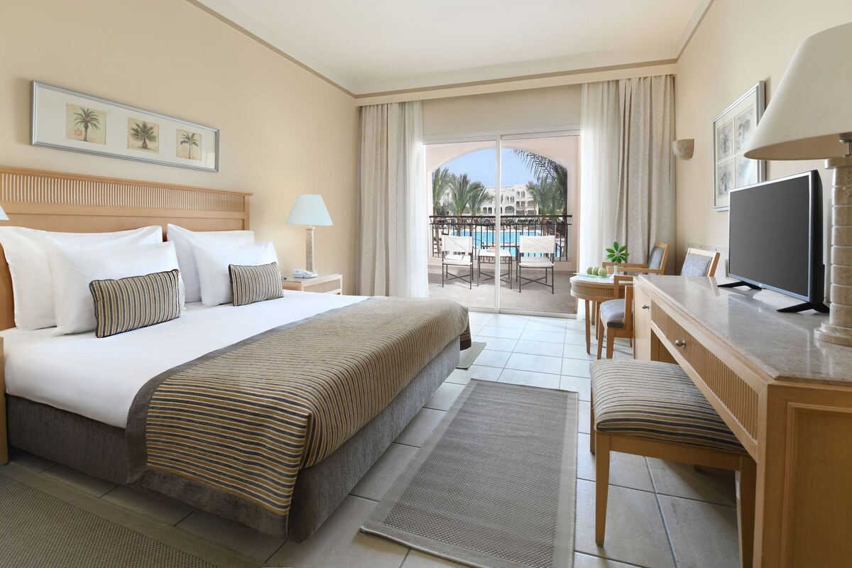 A hotel room with a bed, desk, TV, and a balcony with a pool view.