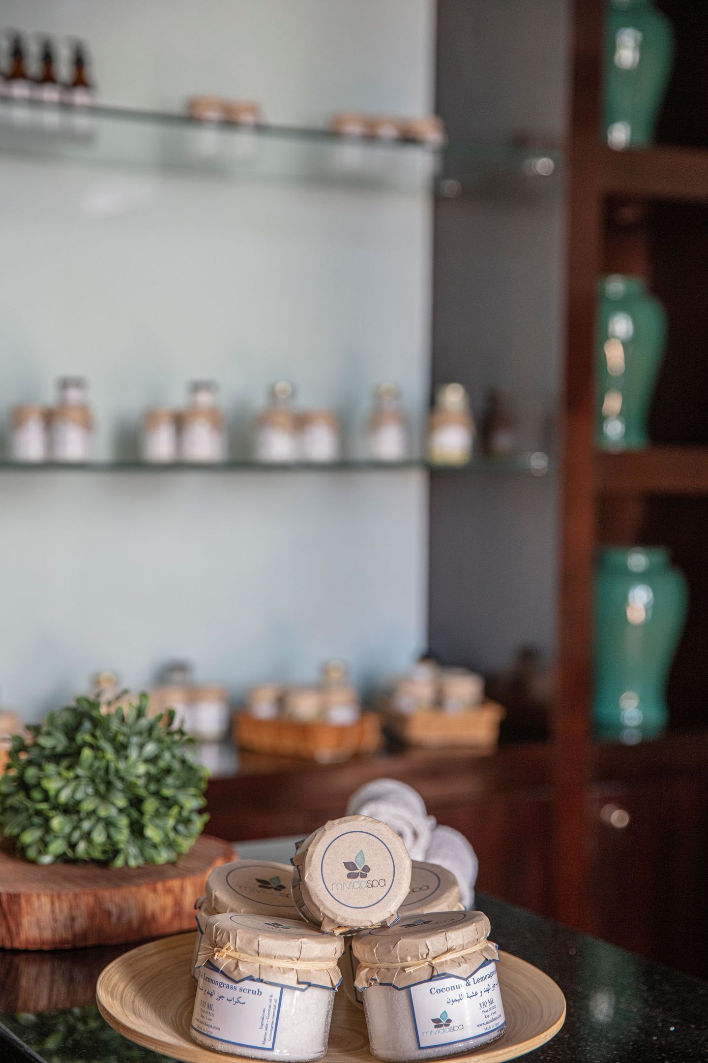 A display of jars with labels on a wooden surface, a green plant, and shelves with jars in the background.