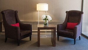 Two armchairs with red cushions, a small table with a vase of white flowers, and a floor lamp in a room with a radiator and a plain wall. - Hotel Universal (Santiago de Compostela)