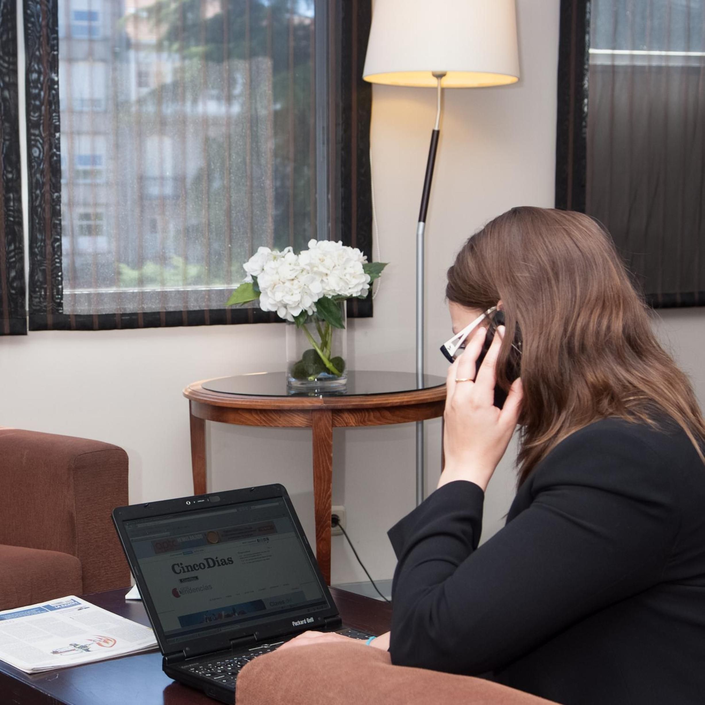 A person seated at a desk with a laptop displaying a news website, a lamp, and a vase with flowers on a small table.