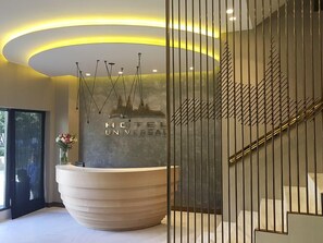 A hotel reception area with a modern design, featuring a curved reception desk, a decorative wall with hanging lights, and a staircase with a metal railing. - Hotel Universal (Santiago de Compostela)