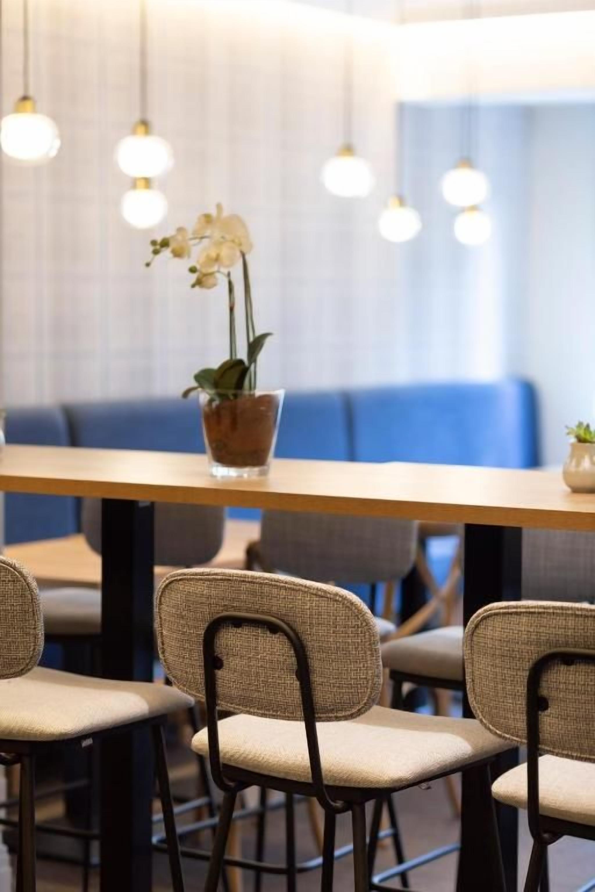 Interior de restaurante con una mesa larga, sillas de mimbre, lámparas colgantes y una orquídea en maceta.