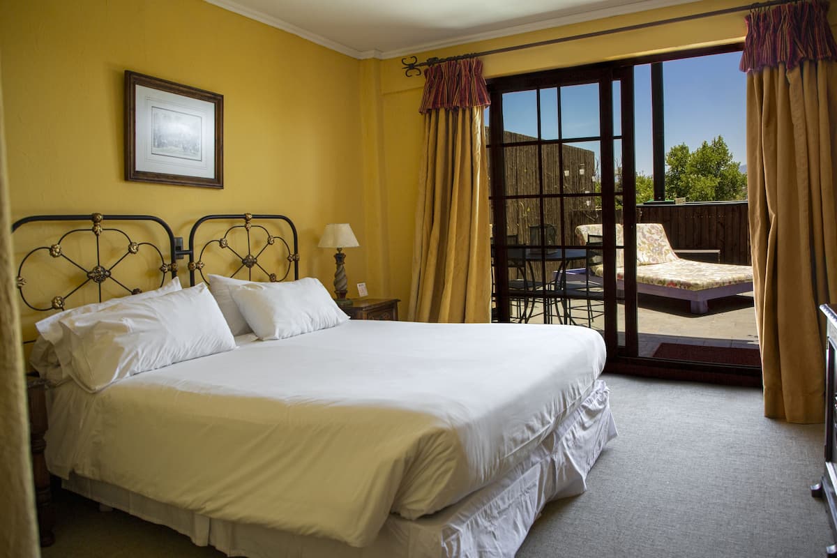 A hotel room with a large bed, a nightstand, a lamp, and a view of the outdoors through a sliding glass door.
