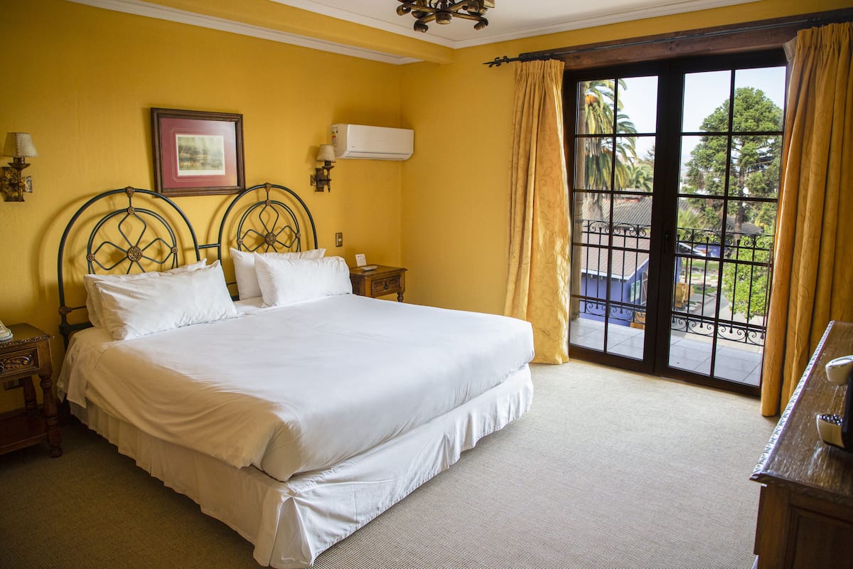 A large bed in a room with yellow walls, a window with a view of the outside, and a nightstand with a lamp.