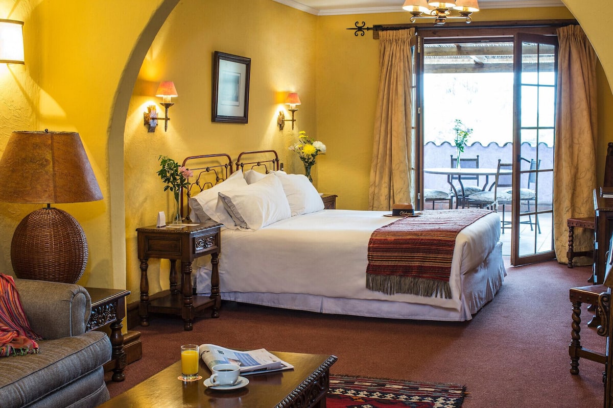 A hotel room with a bed, a sofa, a coffee table, a reading area, and a balcony with a table and chairs.