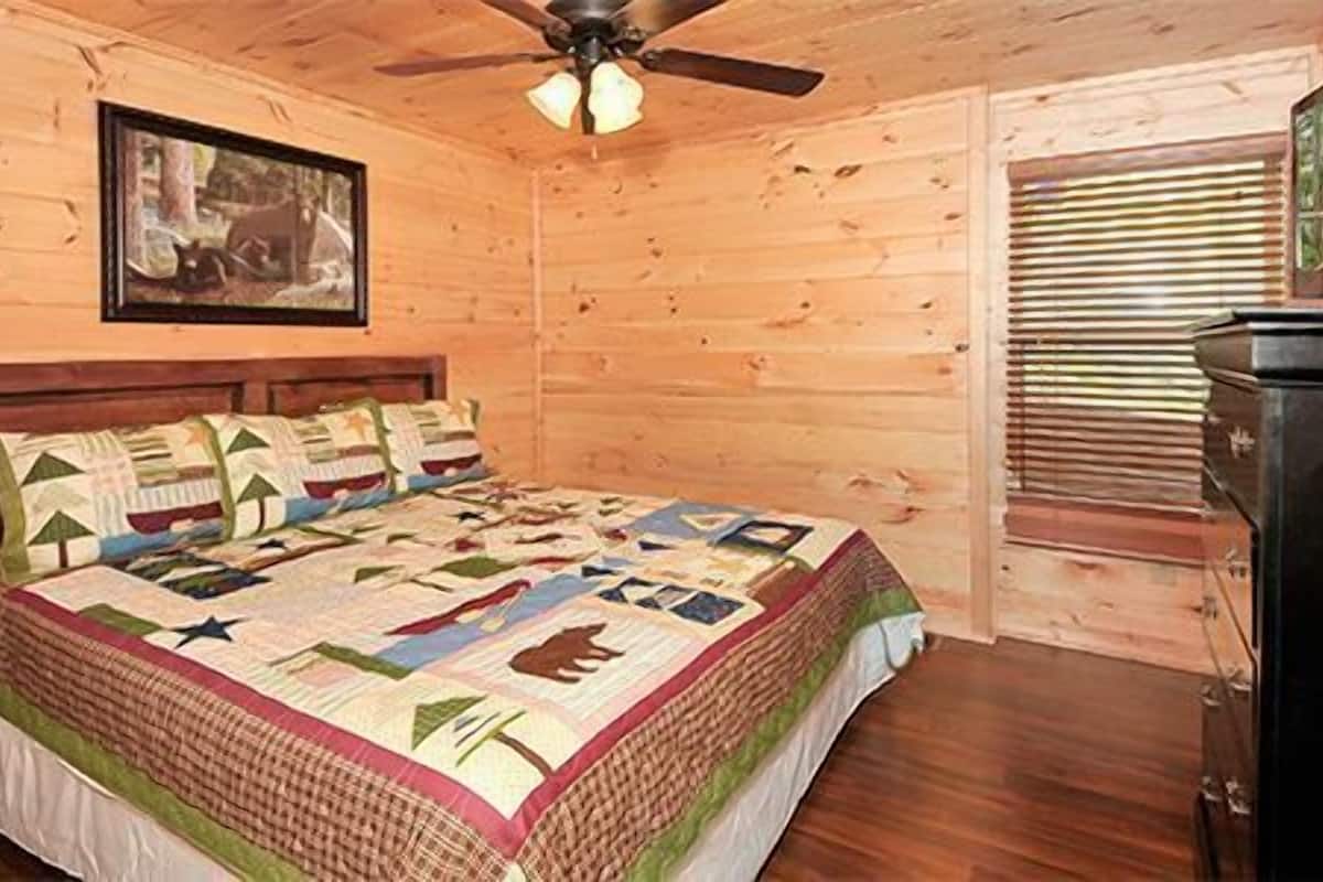 Un dormitorio de cabaña de madera con una cama, un ventilador de techo, un cuadro en la pared y un televisor sobre un aparador.