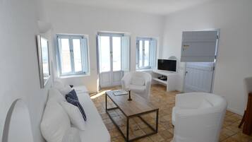 A modern, well-lit living room with white furniture, large windows, and a flat-screen TV.