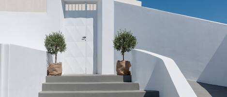 Un escalier blanc mène à une porte ornée d’un motif géométrique, de part et d’autre de laquelle se trouvent deux plantes en pot.