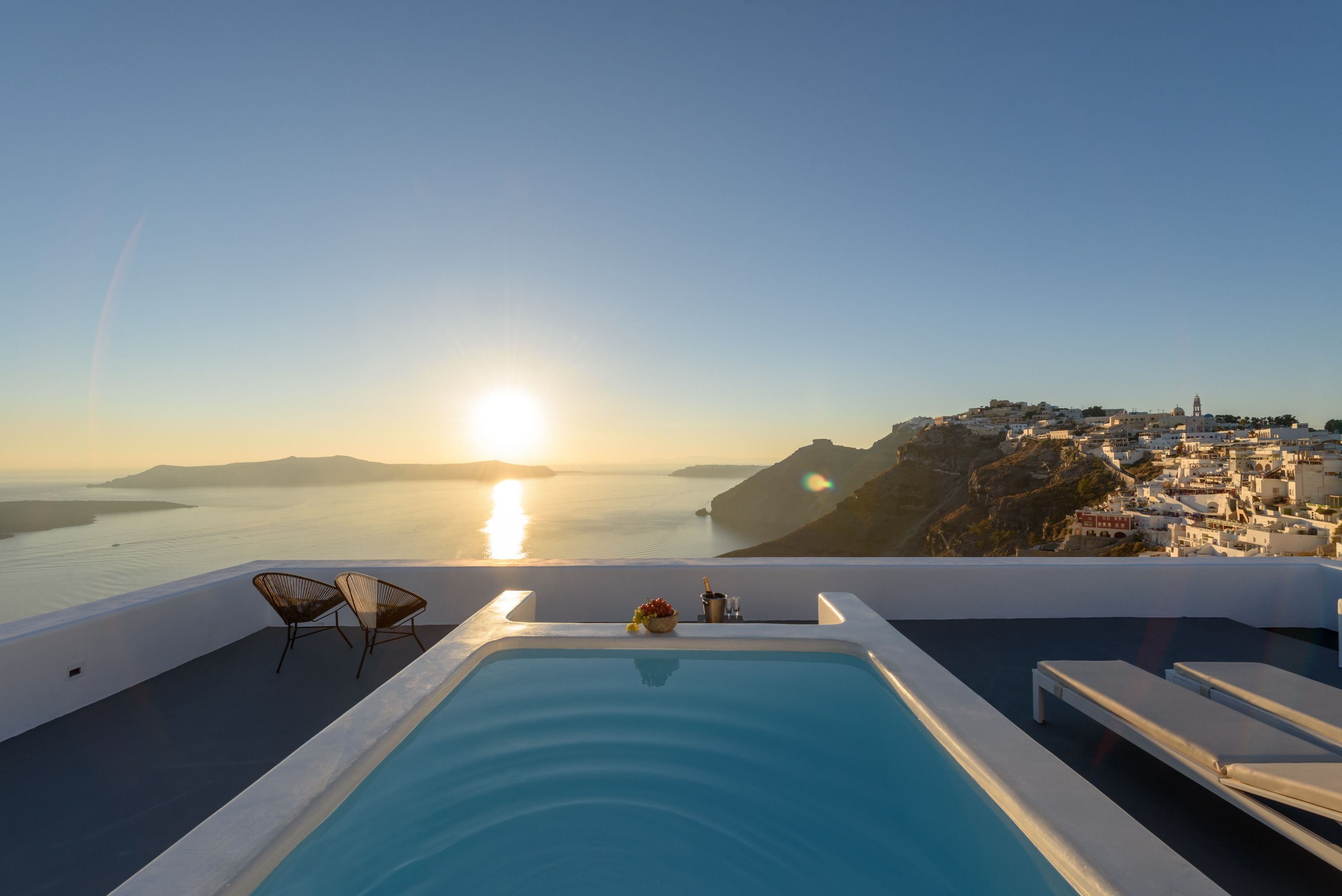 Una piscina infinita en la azotea con sillones de descanso y vistas a un pueblo costero al atardecer.
