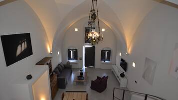 A spacious living area with a high arched ceiling, a chandelier, a dining table with fruit, a sofa, and a staircase.