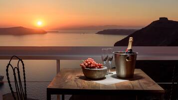 A table with a bottle of champagne, a bowl of grapes, and two wine glasses on a balcony overlooking a sunset over the sea.