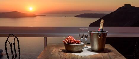 A table with a bottle of champagne, a bowl of grapes, and two wine glasses on a balcony overlooking a sunset over the sea.