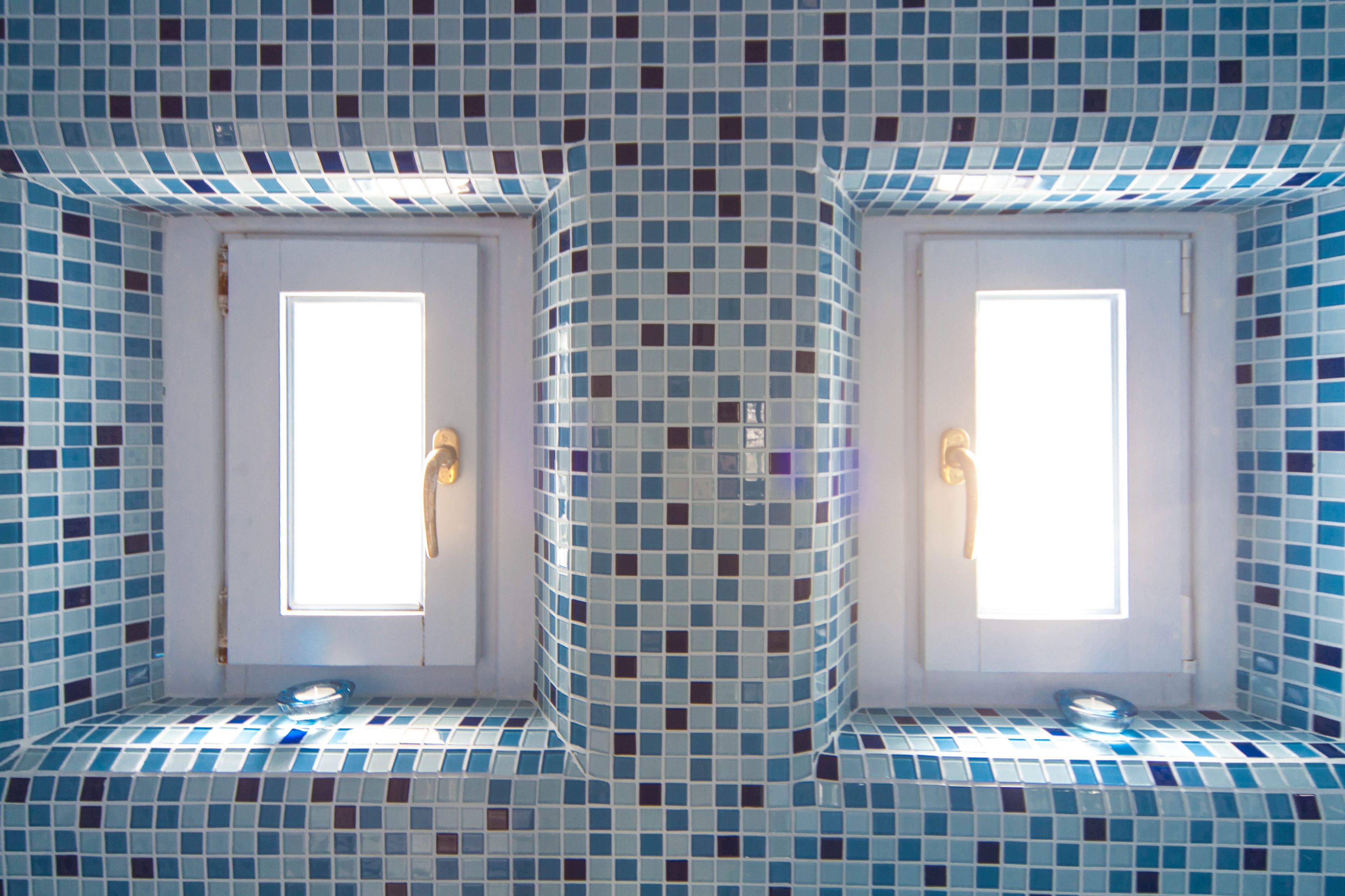 A bathroom with a tiled mosaic floor and walls, two windows with handles, and a light fixture above each window.