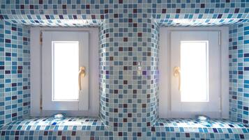 A bathroom with a tiled mosaic floor and walls, two windows with handles, and a light fixture above each window.