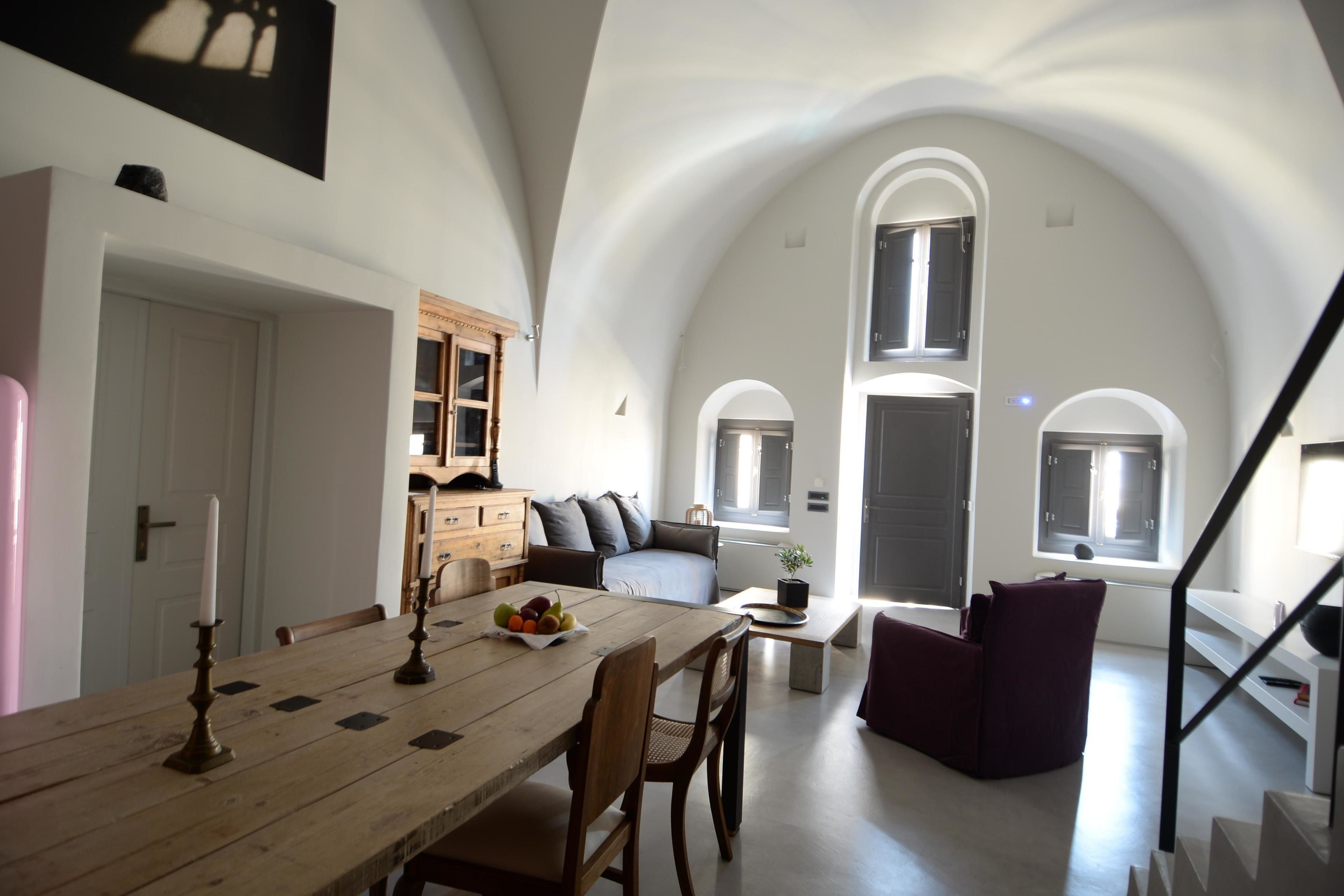 A spacious dining area with a wooden table, chairs, and a fruit bowl. There is a sofa and a small table with a plant. The room has high ceilings with arched windows and a staircase.