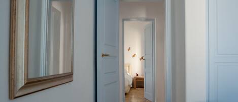 A hallway with a mirror on the left, a bed with a nightstand in the background, and a door on the right.