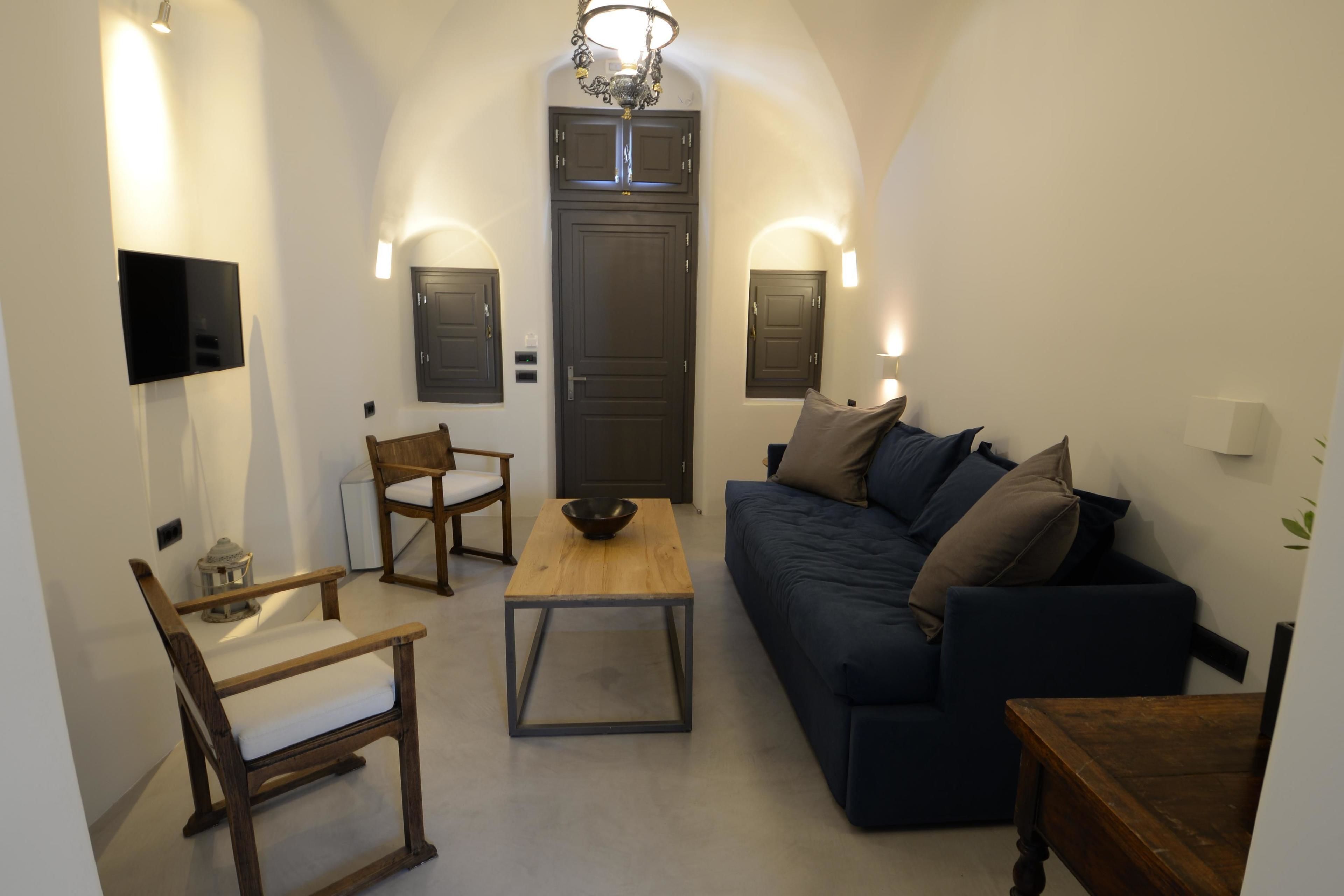 A living room with a dark blue sofa, a wooden coffee table, a wooden chair, a TV mounted on the wall, and a door with two wall-mounted lights.