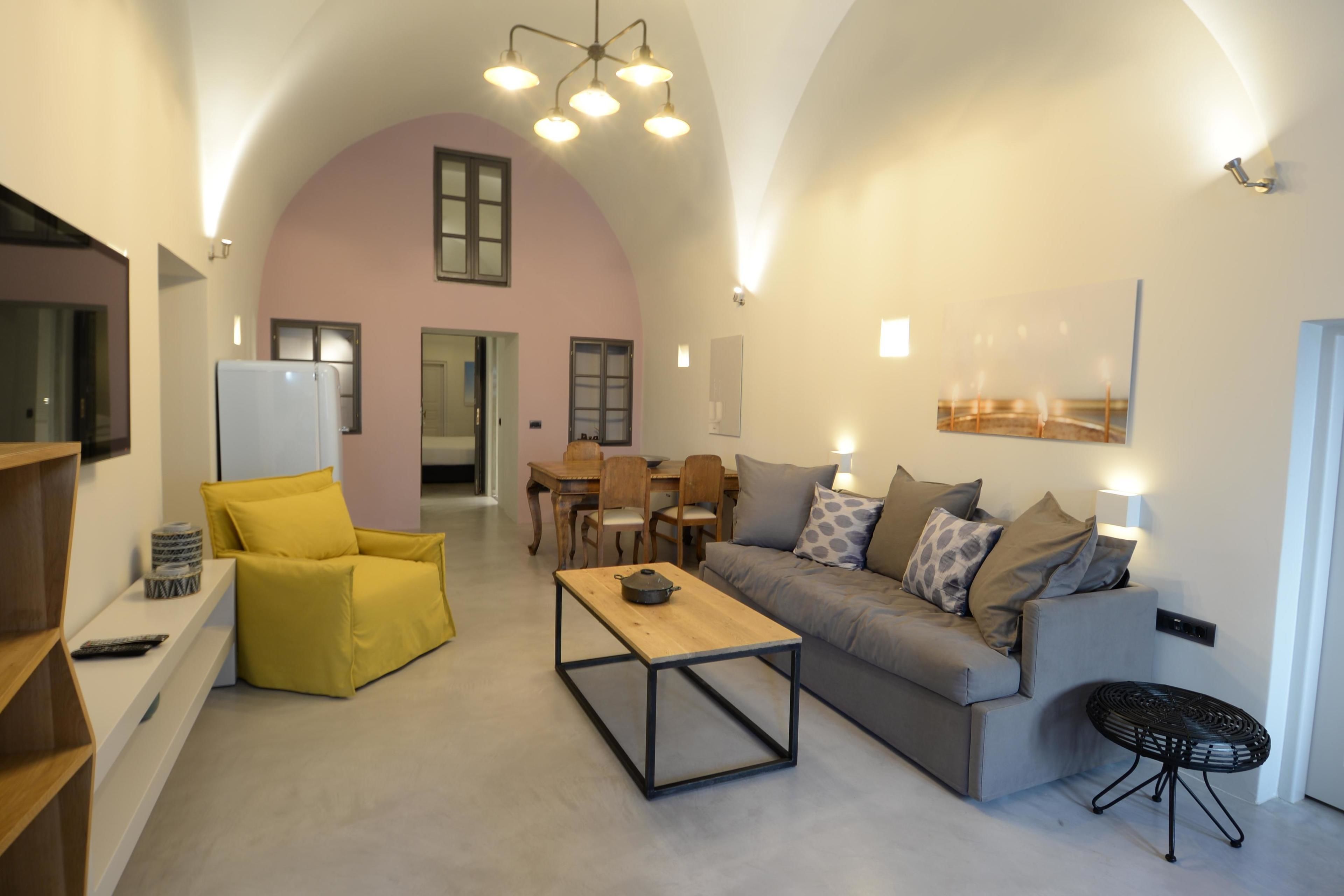 Una sala moderna con un sofá, un sillón amarillo y una mesa de centro. Hay una zona de comedor con una mesa y sillas, y una cocina con un refrigerador.