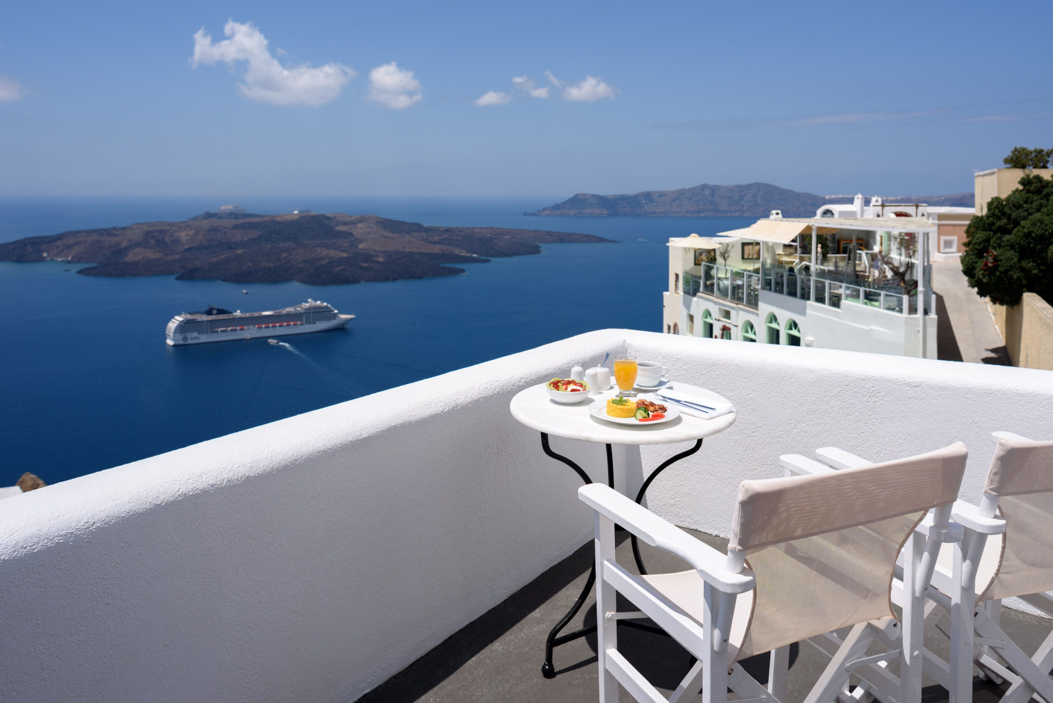 A balcony with a table set for breakfast, overlooking a sea with a cruise ship and distant islands.