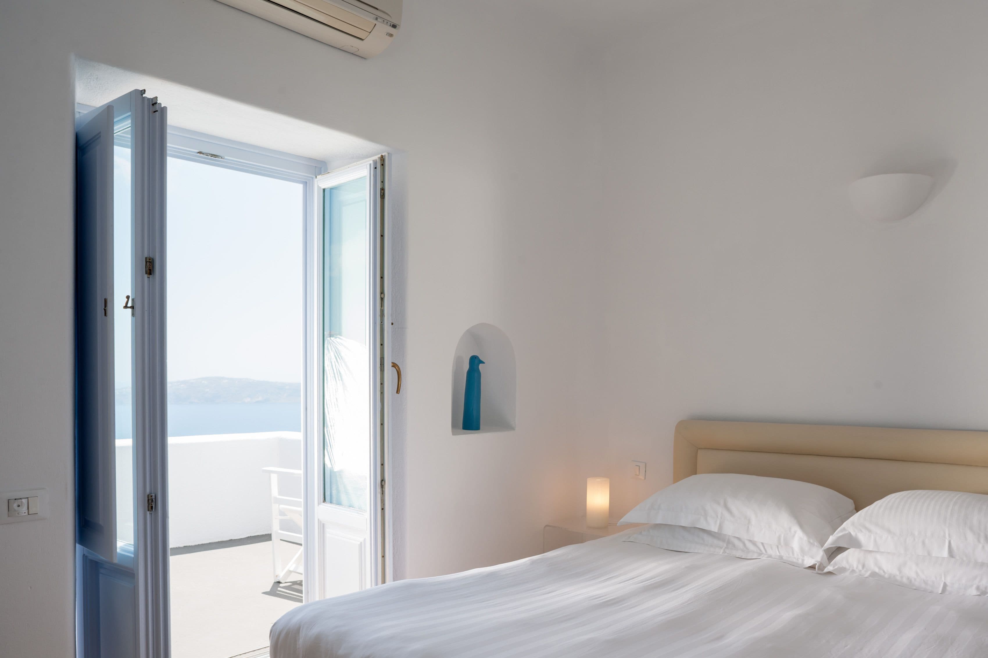 A bedroom with a bed, a nightstand, a wall-mounted light, and a view of the sea through an open door.