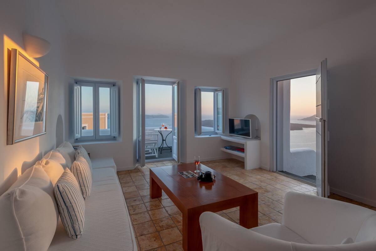 A modern living room with a sofa, a coffee table, and a TV. There are large windows providing views of the outside.