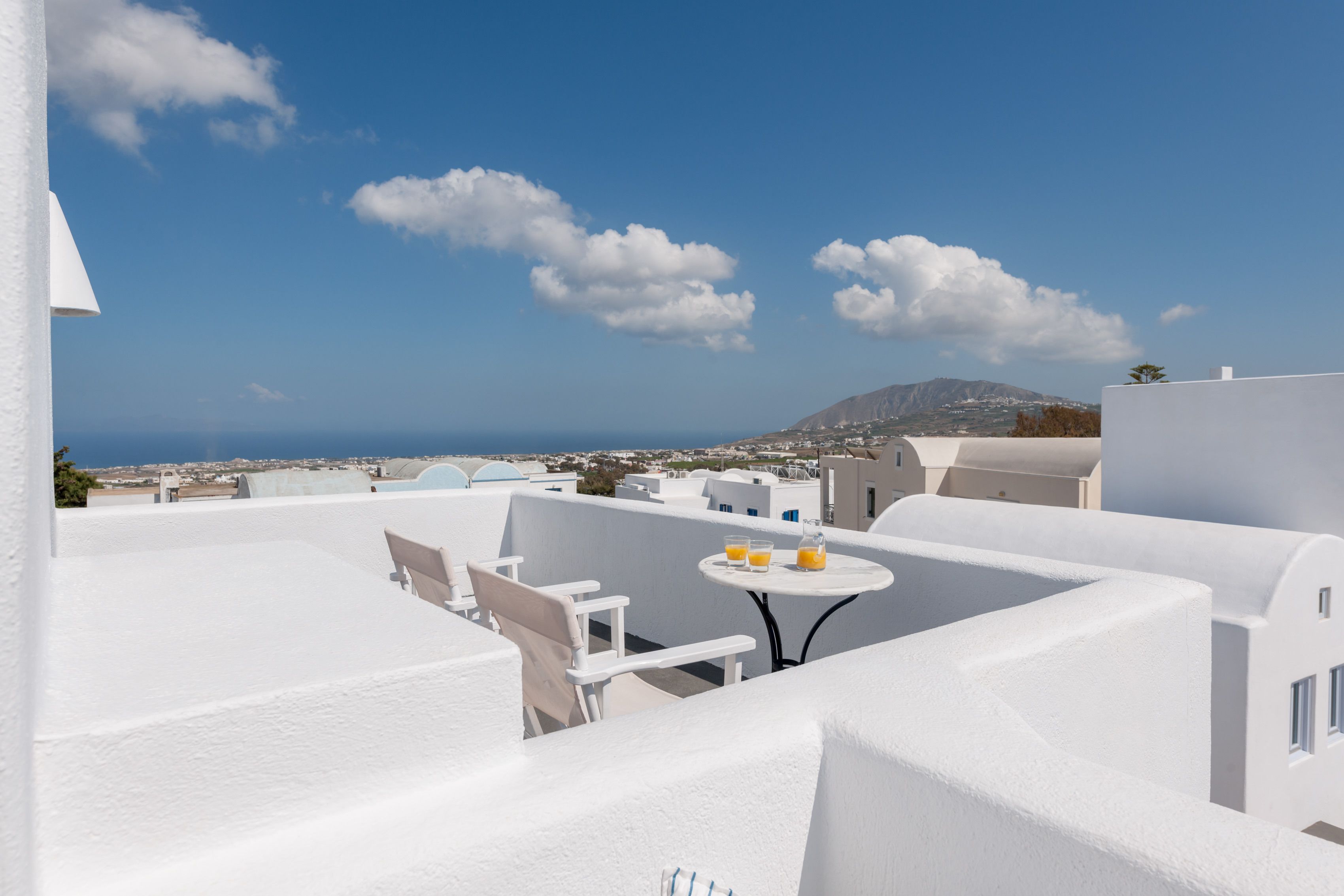 A rooftop terrace with white walls, a round table with two chairs, and a view of the sea and distant buildings.