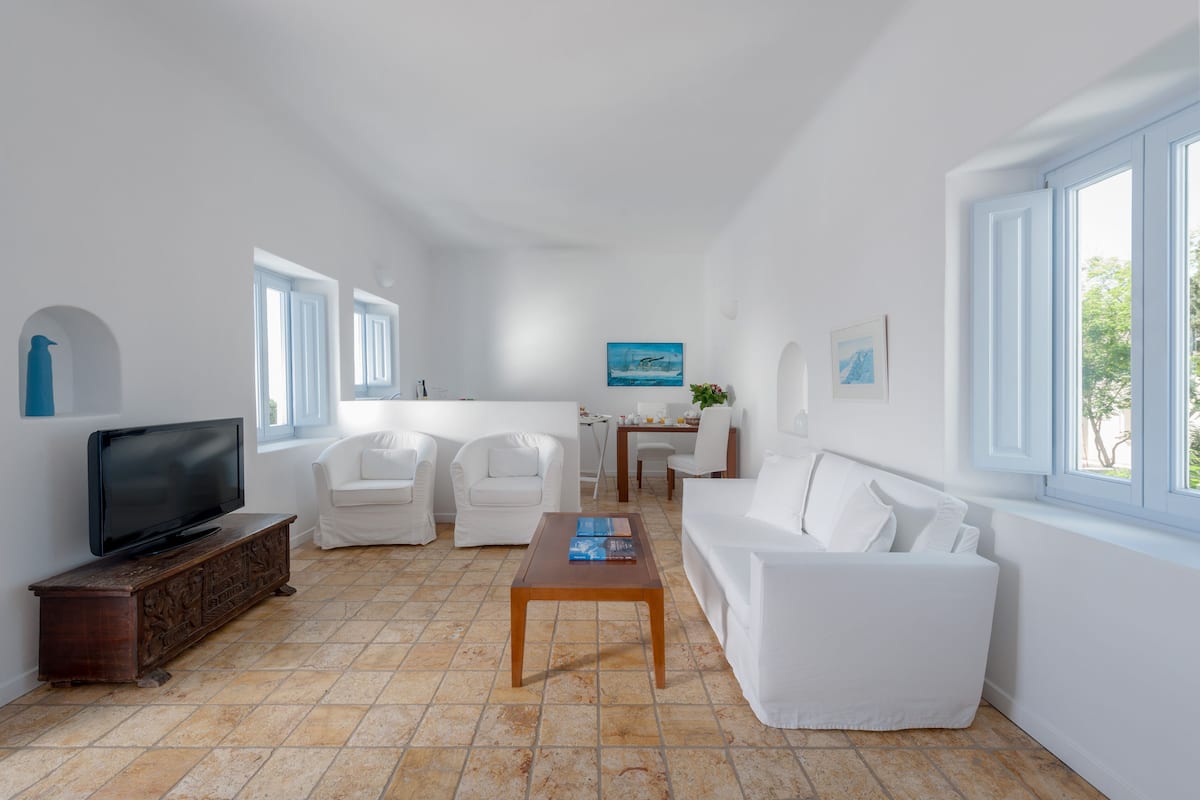 A spacious living area with a television on a wooden cabinet, white furniture, a dining table, and a large window providing natural light.