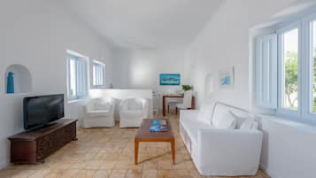 A spacious living area with a television on a wooden cabinet, white furniture, a dining table, and a large window providing natural light.