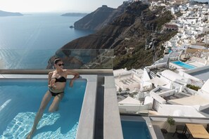 A woman relaxing in a swimming pool with a view of a coastal town and cliffs. - Aria Suites (Santorini)